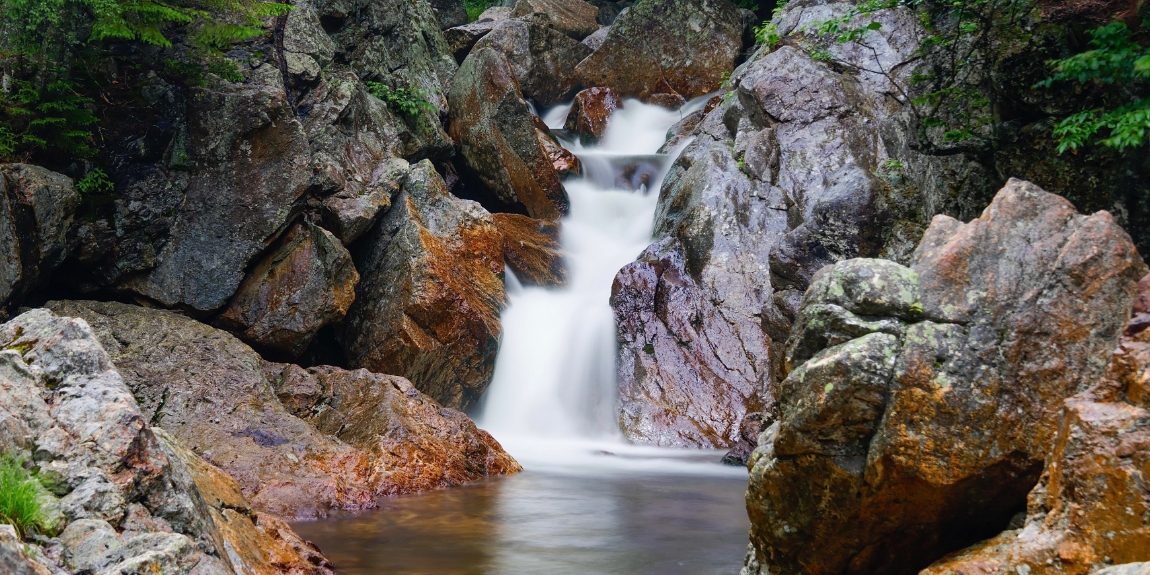 A scenic waterfall cascades down rocky mountains, flowing into a clear stream surrounded by lush greenery.